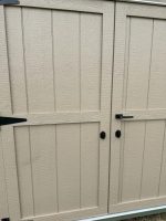 A close-up view of two light tan shed doors with a textured, wood-grain like surface. The doors have black hardware, including hinges on the left door and a latch and handle on the right door, suggesting they open outward. A seam runs vertically between the two doors.