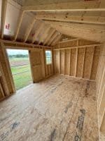 A wide, interior shot from a slightly low angle shows the unfinished wooden framework of a shed or small room. The walls are paneled with OSB (Oriented Strand Board) and feature exposed wooden studs. The ceiling has visible rafters, with some insulation peeking through. On the left wall, an open doorway reveals a view of green grass and an outdoor field, while a small window is visible on the adjacent wall. The floor is covered with OSB panels, some displaying manufacturer markings. The space is empty, showcasing the raw construction materials before finishing.