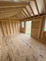 A low-angle interior shot reveals the unfinished wooden framing of a shed or small building. The walls are constructed with OSB (Oriented Strand Board) panels and exposed wooden studs. The ceiling features exposed rafters and insulation. On the far wall, a small window is visible, and in the center, a wooden door stands ajar, offering a glimpse of green grass and daylight outside. The floor is also made of OSB panels, some of which bear printed text and logos. The space appears empty, awaiting further construction and finishing.