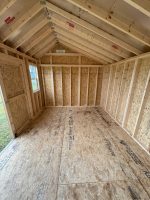 An empty shed under construction, viewed from the open double doors. The floor is made of plywood sheets, and the walls and ceiling are framed with light-colored lumber. A small window is visible on the far wall. The shed appears to be newly built, with no insulation or finishing materials yet installed.