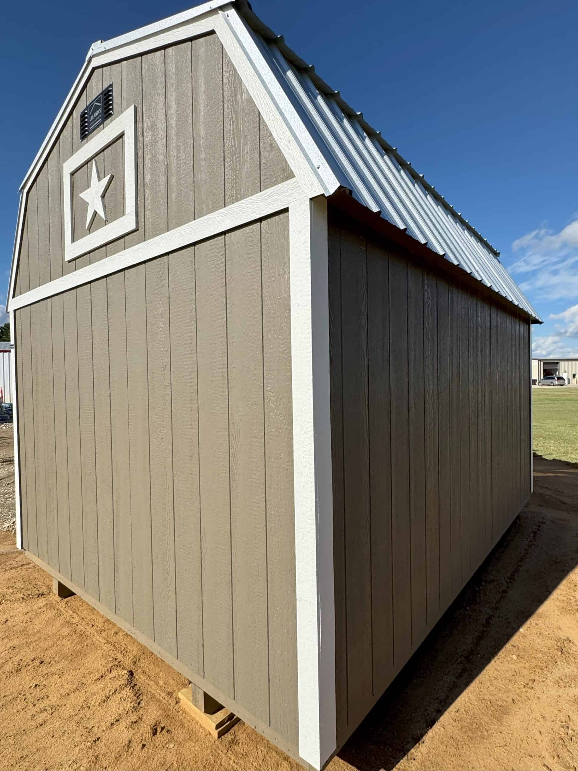 A low-angle exterior shot of a tan barn-style shed, featuring a gambrel roof with a shiny metal surface. The front of the shed has a small white decorative frame with a white star inside. White trim accents the corners and roofline, contrasting with the tan siding. The shed sits on a dirt surface under a bright blue sky with scattered clouds. In the background, a green field and another building are faintly visible.
