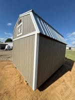 A low-angle exterior shot of a tan barn-style shed, featuring a gambrel roof with a shiny metal surface. The front of the shed has a small white decorative frame with a white star inside. White trim accents the corners and roofline, contrasting with the tan siding. The shed sits on a dirt surface under a bright blue sky with scattered clouds. In the background, a green field and another building are faintly visible.