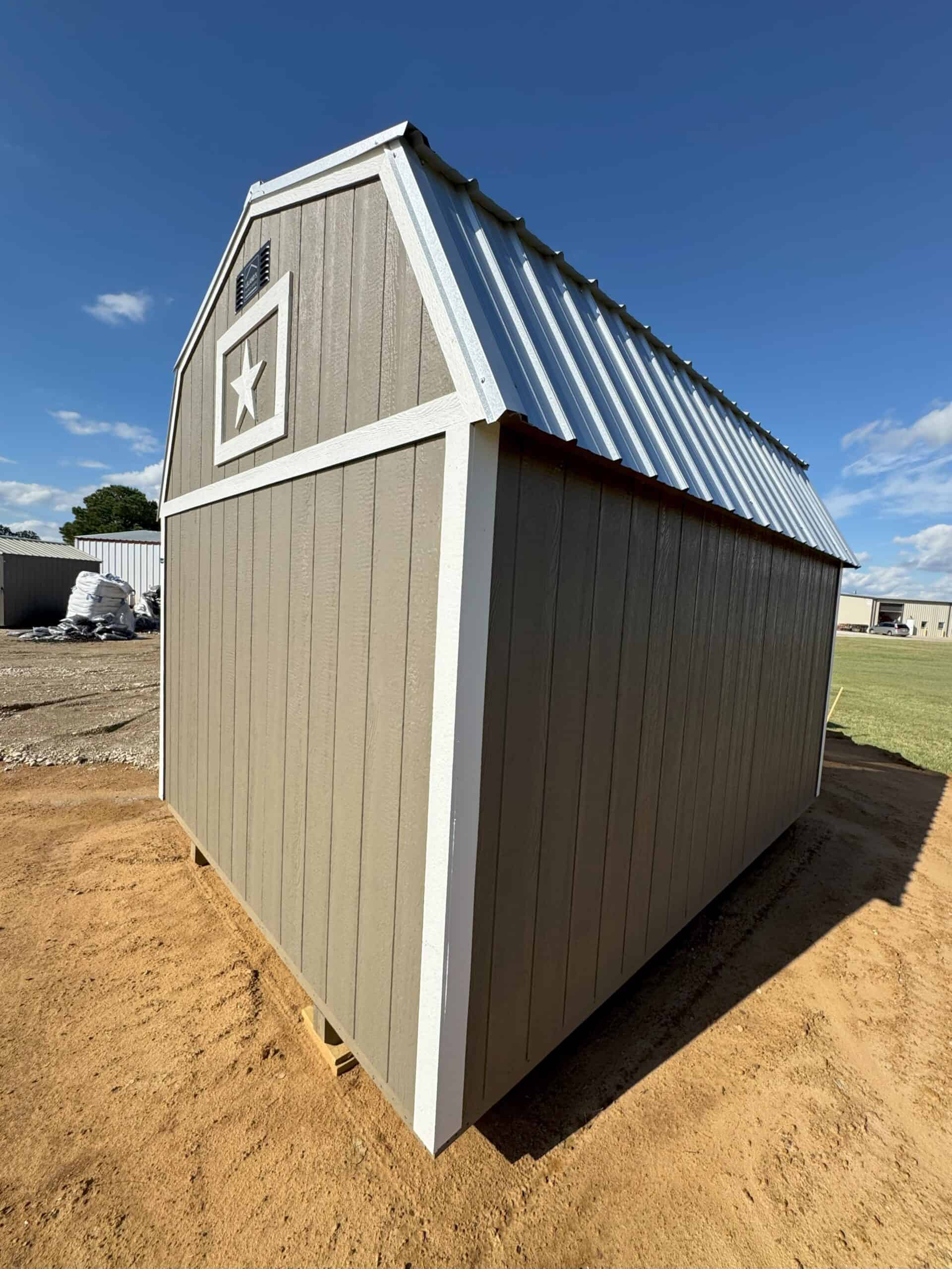A low-angle exterior shot of a tan barn-style shed, featuring a gambrel roof with a shiny metal surface. The front of the shed has a small white decorative frame with a white star inside. White trim accents the corners and roofline, contrasting with the tan siding. The shed sits on a dirt surface under a bright blue sky with scattered clouds. In the background, a green field and another building are faintly visible.