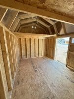 An interior view of an unfinished shed, showcasing its wooden frame and plywood floor. The walls are framed with vertical studs, and the ceiling has exposed rafters leading up to a peak. There's an open doorway on the right, revealing a gravel ground and hints of buildings outside. A small vent is visible high on the back wall.