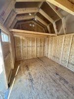 An indoor shot of an empty wooden shed reveals unpainted walls made of vertical oriented strand boards and a floor of the same material. The shed has an open, unfinished ceiling with exposed wooden beams and rafters, indicating the structure of a gambrel roof. A small window is visible on the left wall, allowing natural light to enter.