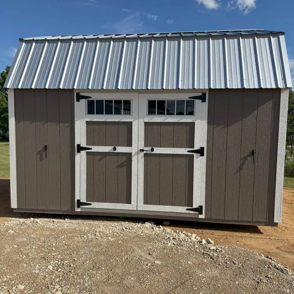 A frontal exterior shot of a shed with a gambrel roof made of shiny metal. The shed's body is painted in two tones: the central double doors and their surrounding panels are dark brown, while the outer wall sections on either side are a lighter tan. The double doors feature small windows with white mullions and are accented with black hinges and handles. White trim frames the doors and the roofline. The shed rests on a gravel and dirt surface under a bright blue sky with a few white clouds.