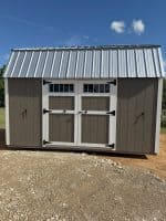 A frontal exterior shot of a shed with a gambrel roof made of shiny metal. The shed's body is painted in two tones: the central double doors and their surrounding panels are dark brown, while the outer wall sections on either side are a lighter tan. The double doors feature small windows with white mullions and are accented with black hinges and handles. White trim frames the doors and the roofline. The shed rests on a gravel and dirt surface under a bright blue sky with a few white clouds.