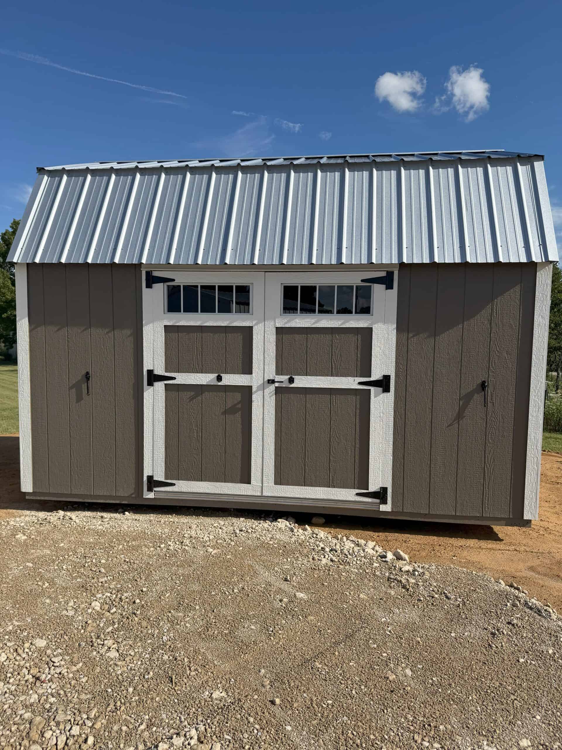 A frontal exterior shot of a shed with a gambrel roof made of shiny metal. The shed's body is painted in two tones: the central double doors and their surrounding panels are dark brown, while the outer wall sections on either side are a lighter tan. The double doors feature small windows with white mullions and are accented with black hinges and handles. White trim frames the doors and the roofline. The shed rests on a gravel and dirt surface under a bright blue sky with a few white clouds.