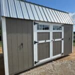 A rectangular shed with dark brown siding and white trim is positioned on a gravel ground under a clear blue sky. It features white-framed double doors with black hinges and handles, topped by two rectangular windows. A corrugated metal roof caps the structure, and two small black handles are visible on the far left and right sides of the shed.