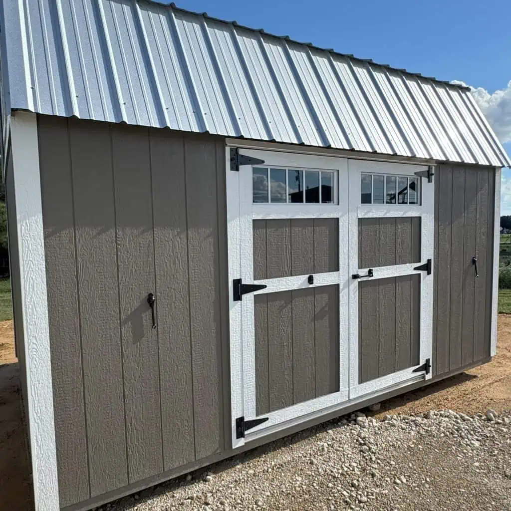 A rectangular shed with dark brown siding and white trim is positioned on a gravel ground under a clear blue sky. It features white-framed double doors with black hinges and handles, topped by two rectangular windows. A corrugated metal roof caps the structure, and two small black handles are visible on the far left and right sides of the shed.