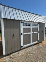 A rectangular shed with dark brown siding and white trim is positioned on a gravel ground under a clear blue sky. It features white-framed double doors with black hinges and handles, topped by two rectangular windows. A corrugated metal roof caps the structure, and two small black handles are visible on the far left and right sides of the shed.