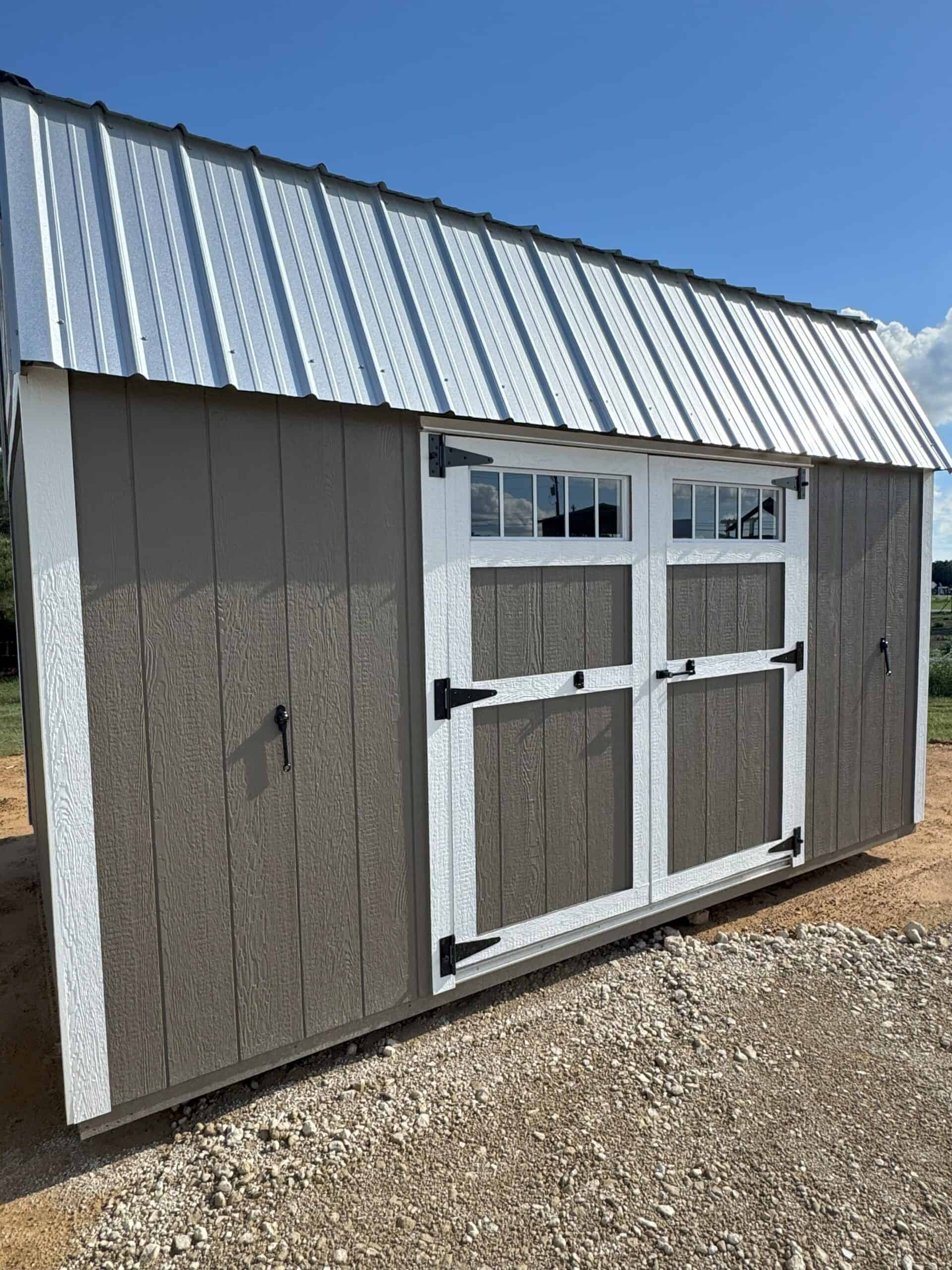 A rectangular shed with dark brown siding and white trim is positioned on a gravel ground under a clear blue sky. It features white-framed double doors with black hinges and handles, topped by two rectangular windows. A corrugated metal roof caps the structure, and two small black handles are visible on the far left and right sides of the shed.