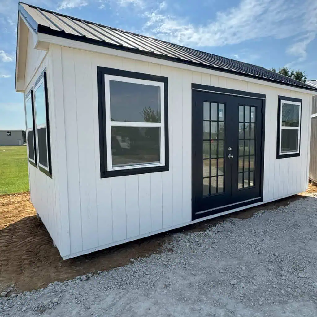 A small, white shed-like building with a dark metal roof, black-framed double doors, and several black-framed windows, sitting on gravel and dirt under a blue sky with scattered clouds.