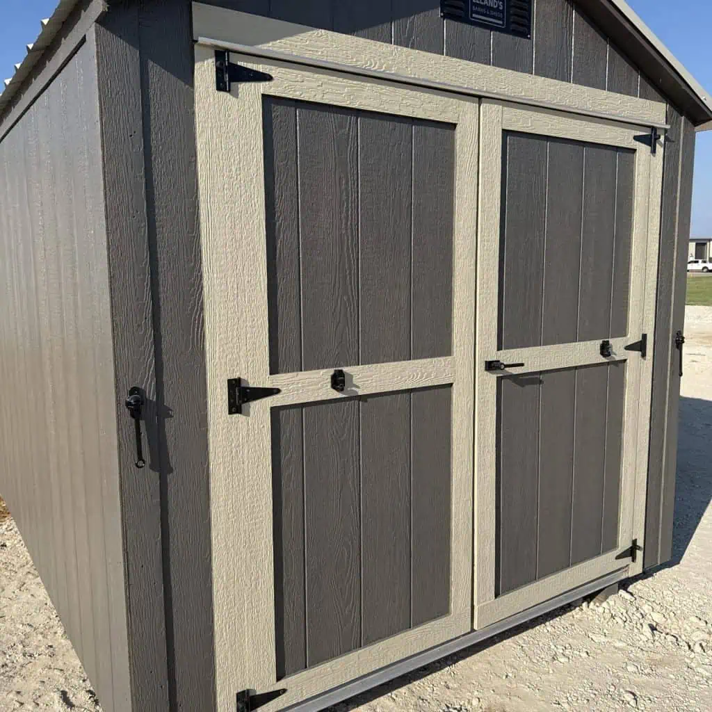 A shed with dark brown siding and light tan trim around the double doors, featuring black hinges and handles, under a clear blue sky. A "LELAND'S" logo is visible at the top of the gable.