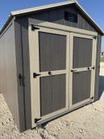 A shed with dark brown siding and light tan trim around the double doors, featuring black hinges and handles, under a clear blue sky. A "LELAND'S" logo is visible at the top of the gable.