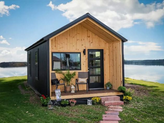A tiny house with black siding and a natural wood front porch area stands on a grassy patch beside a calm body of water. The house has a black roof, a large horizontal window on the left side of the front, and a black front door with glass panes on the right. The small wooden porch is furnished with two chairs, plants in pots, and decorative items. Red brick steps lead from the porch down to a winding brick path on the grass. The background features a wide expanse of water reflecting the blue sky and scattered white clouds.