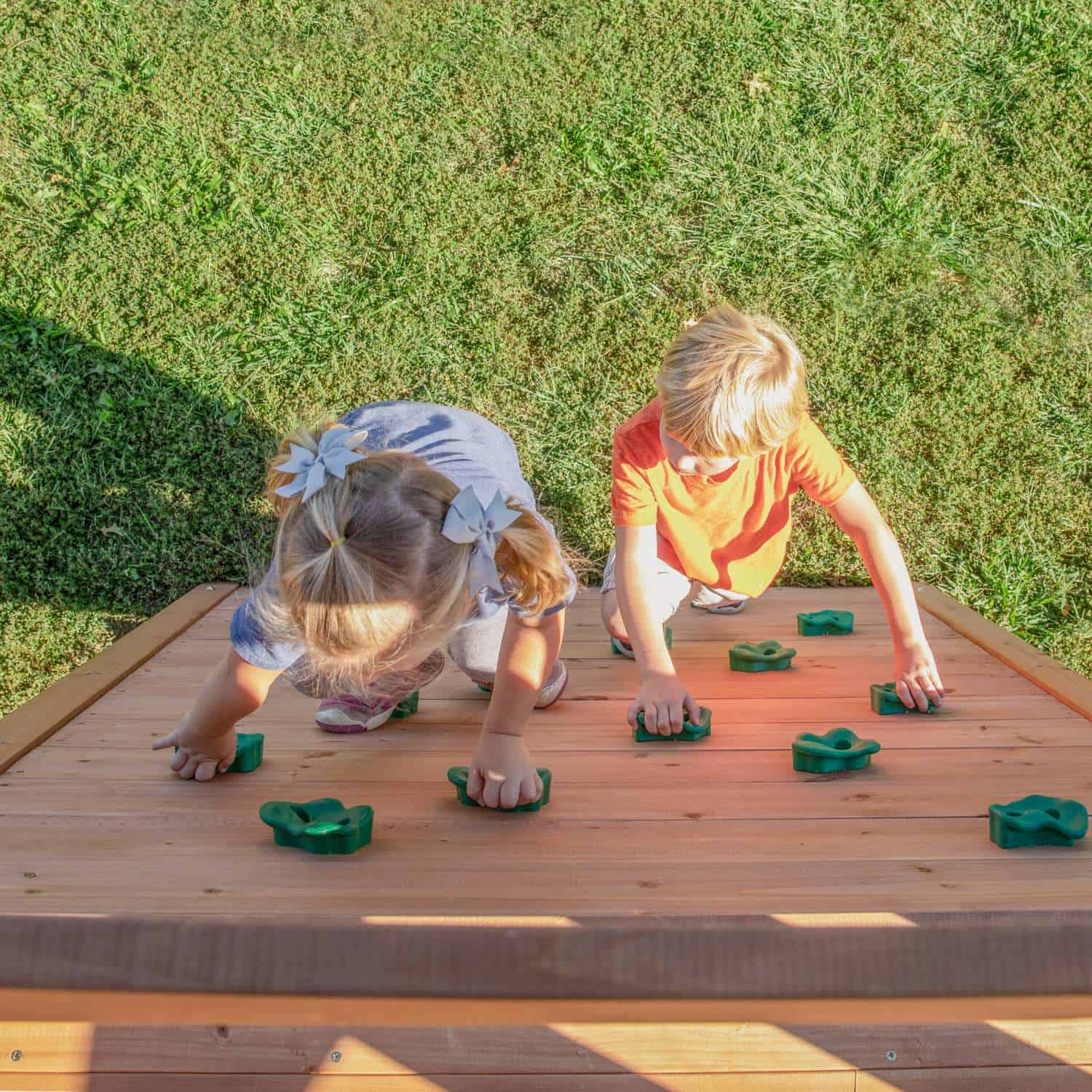Two young children are seen from above, crawling on a wooden climbing wall with green handholds. The girl on the left has blonde hair tied in two pigtails with white bows and is wearing a light-colored top. The boy on the right has blonde hair and is wearing an orange t-shirt and khaki shorts. They are both reaching for the handholds, and green grass is visible in the background.