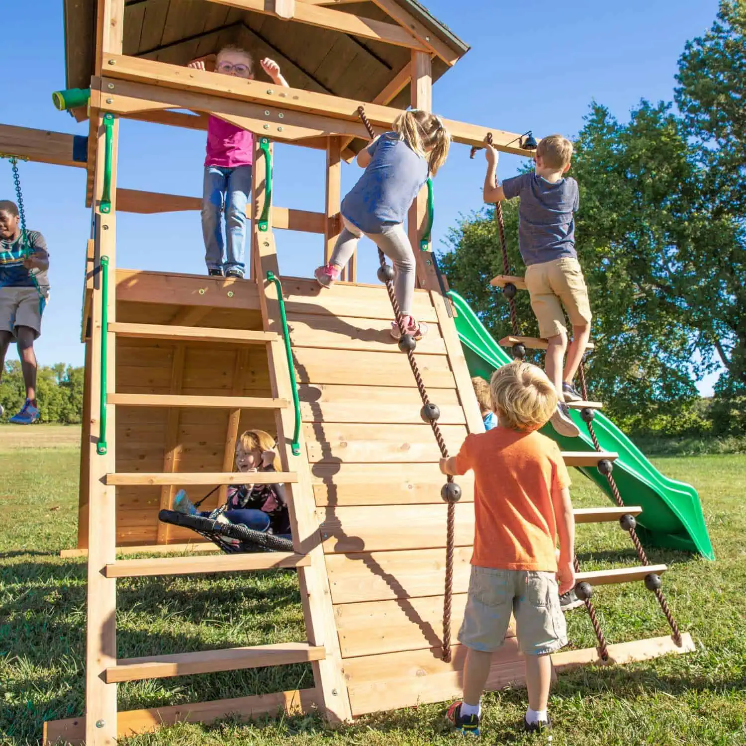 A group of children are actively playing on a wooden playground structure under a clear blue sky. In the foreground, a boy in an orange shirt and khaki shorts is climbing a rope ladder with wooden steps. Below him, two younger children are sitting in a net swing. To the left, another child is seen climbing up a ramp with rope and spherical grips, while a green slide is visible on the right side of the playset.