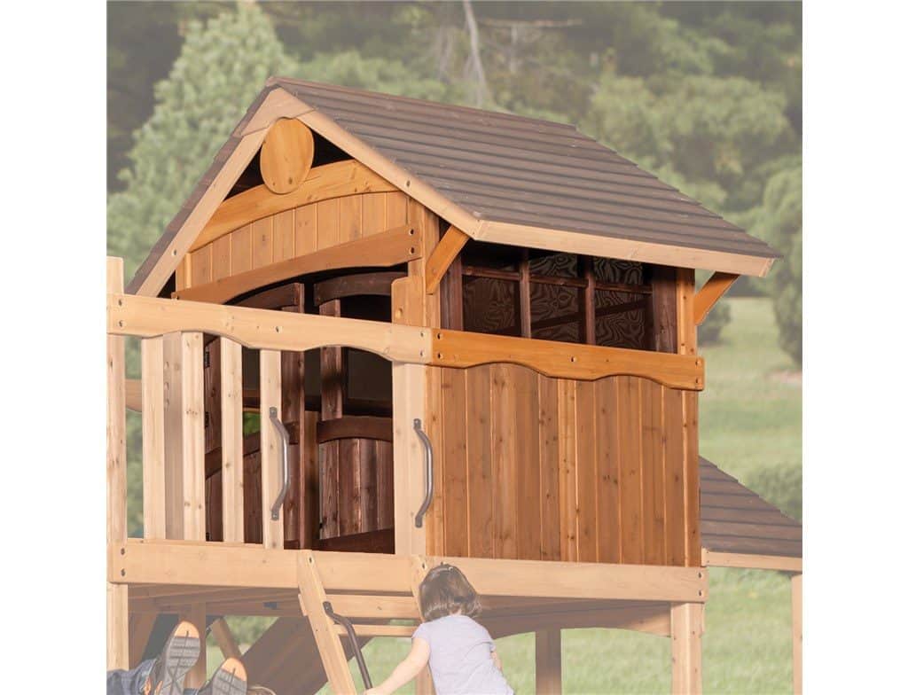 A low-angle shot shows a wooden playhouse and a small girl climbing a ladder. The playhouse has a brown roof, wood-paneled walls, and a small window. It is built on a raised platform, part of a larger playset, with a wooden railing in the foreground. The girl, seen from behind, wears a light purple top and is ascending a ladder made of wooden rungs. In the background, out-of-focus trees and green foliage suggest an outdoor setting. The image has a light, natural feel.