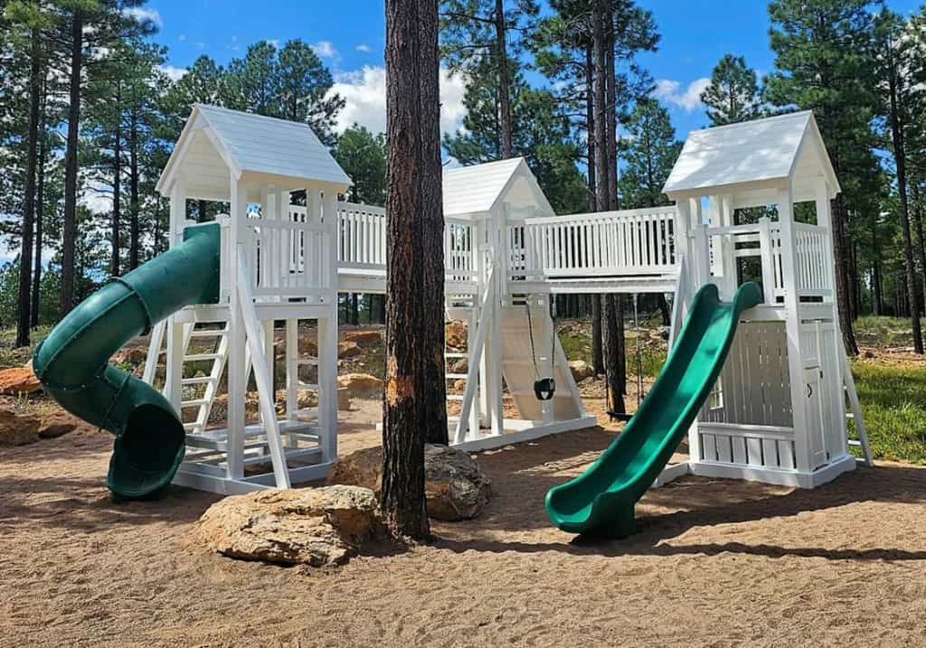 A bright white wooden vinyl playset with two green slides is set in a natural, rustic environment with tall pine trees. The playset features multiple towers with peaked roofs connected by a bridge, and includes various ladders and swings. The ground is a mix of sand, dirt, and rocks, with a dense forest in the background under a partly cloudy sky.