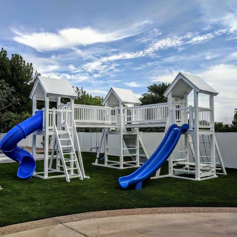 A white wooden vinyl playground set with blue slides stands on a vibrant green turf under a partly cloudy sky. The playset features multiple towers with peaked roofs, connected by bridges, and includes various ladders, a tube slide on the left, and an open slide on the right. Trees and a white fence are visible in the background.