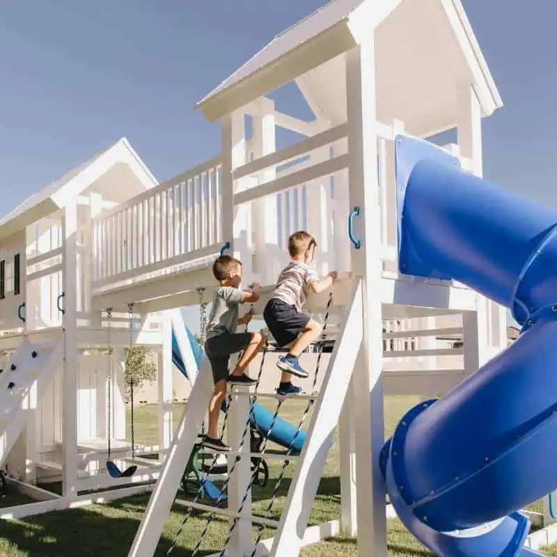 Two young boys are actively playing on a bright white outdoor vinyl playset with blue accents. One boy, wearing a grey t-shirt and shorts, is climbing a rope ladder, while the other, in a striped t-shirt and blue shorts, is ascending a wooden ladder next to him. A blue tube slide is prominent on the right.