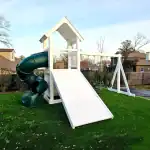 White vinyl playset with a covered tower, ladder, and a long white slide, set on a green artificial turf yard.