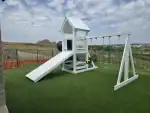White vinyl playset with a peaked roof, swings, and a green slide set on a grassy yard.