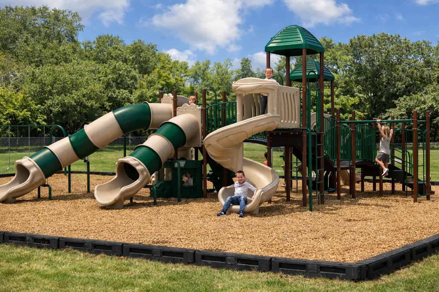 Gemini said A group of children play on a large playground structure featuring a neutral color palette of tan and forest green. The play set is situated on a bed of light brown wood mulch contained by a black plastic border within a grassy field. The equipment includes two long, striped tube slides in tan and green, as well as a tan spiral slide where a young boy is seated at the base. Other children are visible on the elevated platforms and hanging from a set of overhead monkey bars. The structure is accented with green gabled roofs on the tower sections, and a dense line of leafy green trees fills the background under a blue sky with scattered white clouds.