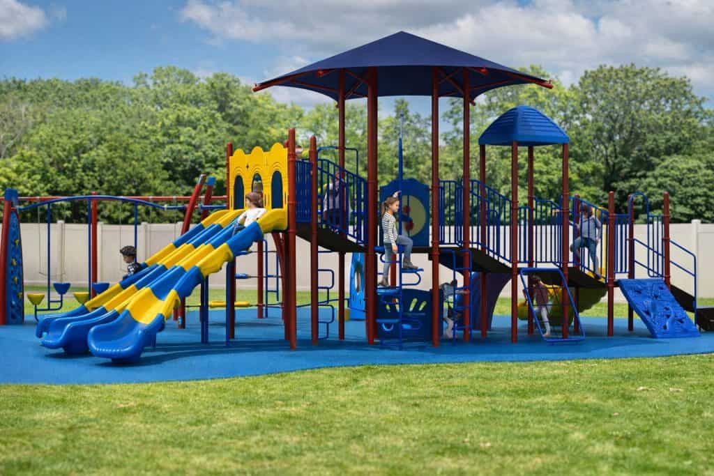 Gemini said A vibrant, multi-colored playground structure featuring shades of blue, red, and yellow sits on a bright blue safety surface surrounded by green grass. Several children are actively playing on the equipment, with one child sliding down a triple-track blue and yellow slide, another climbing a blue spiral ladder, and others positioned on the various elevated platforms. The central tower is covered by a large, dark blue conical shade roof, while a smaller blue domed roof sits atop an adjacent section. The background shows a light-colored fence and a line of dense green trees under a bright, partly cloudy sky.