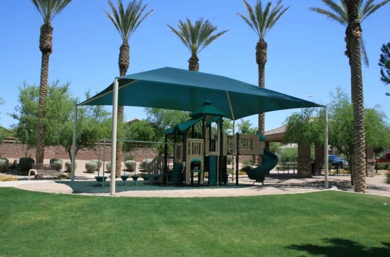 A tan and dark green playground structure sits shaded beneath a large, rectangular dark green sun sail in a desert park setting. The play set includes a spiral slide, climbing platforms, and a row of stepping pods, all positioned on a sand-colored surface. Tall palm trees and smaller desert trees frame the playground, with a green grassy lawn in the foreground and a covered picnic pavilion in the background under a clear blue sky.