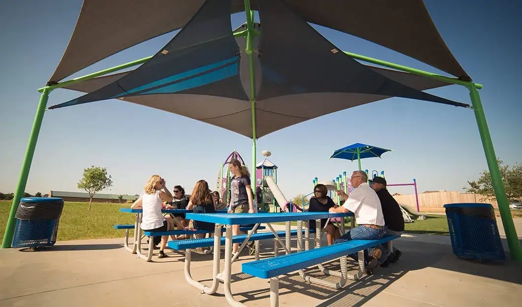 Children and adults enjoy shade under a large outdoor canopy at a playground.