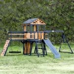 Children's wooden playset with slide, swings, and climbing wall in backyard.