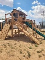 Wooden playhouse with slides and climbing wall in outdoor playground.