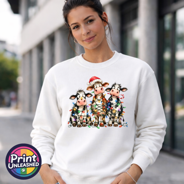 Three wide-eyed Christmas cows tangled in colourful lights, one wearing a Santa hat and another holding a candy cane, printed on a white unisex sweatshirt female model - Print Unleashed