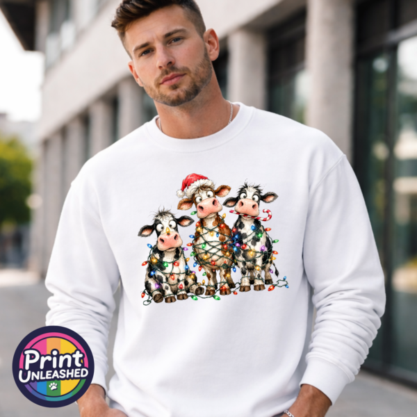 Three wide-eyed Christmas cows tangled in colourful lights, one wearing a Santa hat and another holding a candy cane, printed on a white unisex sweatshirt male model - Print Unleashed