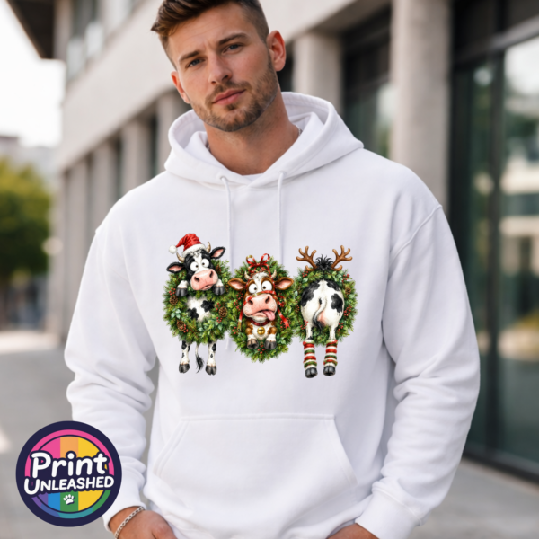 Three Christmas cows in festive wreaths, one wearing a Santa hat and another facing backwards with antlers and striped leg warmers, printed on a white unisex hoodie male model - Print Unleashed