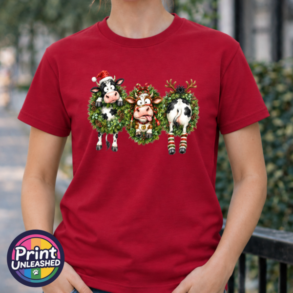 Three Christmas cows in festive wreaths, one wearing a Santa hat and another facing backwards with antlers and striped leg warmers, printed on a cherry red premium unisex T-shirt female model - Print Unleashed