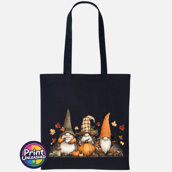 Three autumn gonks in tall pointed hats with pumpkins, a takeaway cup, a black cat and falling leaves printed on a black tote bag - Print Unleashed