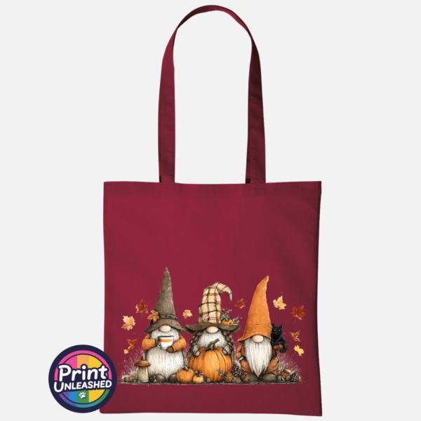 Three autumn gonks in tall pointed hats with pumpkins, a takeaway cup, a black cat and falling leaves printed on a deep red tote bag - Print Unleashed