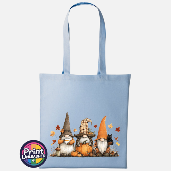 Three autumn gonks in tall pointed hats with pumpkins, a takeaway cup, a black cat and falling leaves printed on a sky blue tote bag - Print Unleashed