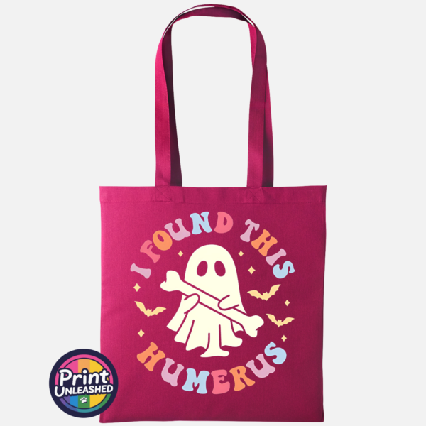 Cute ghost holding a bone beneath colourful I Found This Humerus lettering, surrounded by bats and stars, printed on a cranberry tote bag - Print Unleashed