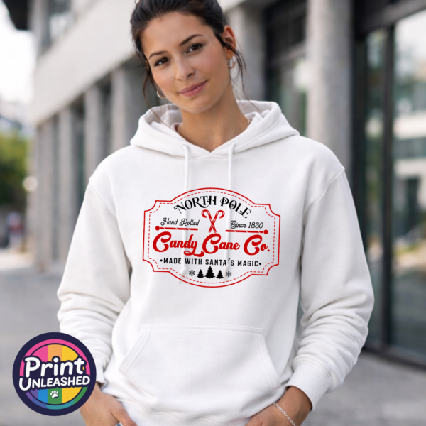 Vintage North Pole Candy Cane Co. design with crossed candy canes, decorative red borders, fir trees and snowflakes, printed on a white unisex hoodie female model - Print Unleashed