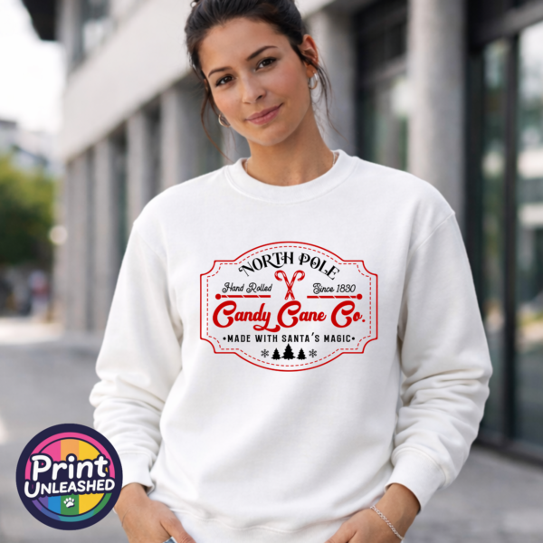 North Pole Candy Cane Co. female sweat Vintage North Pole Candy Cane Co. design with crossed candy canes, decorative red borders, fir trees and snowflakes, printed on a white unisex sweatshirt female model - Print Unleashed
