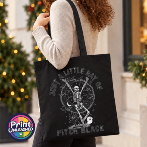 Model image of a black tote bag featuring a dancing skeleton surrounded by bats, stars, a crescent moon, a cat and ghost with Just a Little Ray of Pitch Black lettering - Print Unleashed
