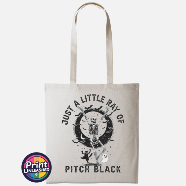 Pitch Black natural Natural tote bag featuring a dancing skeleton surrounded by bats, stars, a crescent moon, a cat and ghost with Just a Little Ray of Pitch Black lettering - Print Unleashed
