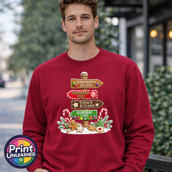 Gingerbread-style signpost pointing to Gingerbread Village, Peppermint Post, Cookie Kitchen and Gumdrop Lane, with sweets and biscuits, printed on a cherry red unisex sweatshirt male model - Print Unleashed