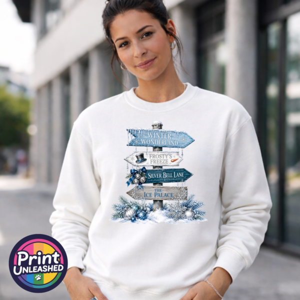 Snow-covered signpost pointing to Winter Wonderland, Frosty’s Freeze, Silver Bell Lane and the Ice Palace, with blue and silver decorations, printed on a white unisex sweatshirt female model - Print Unleashed