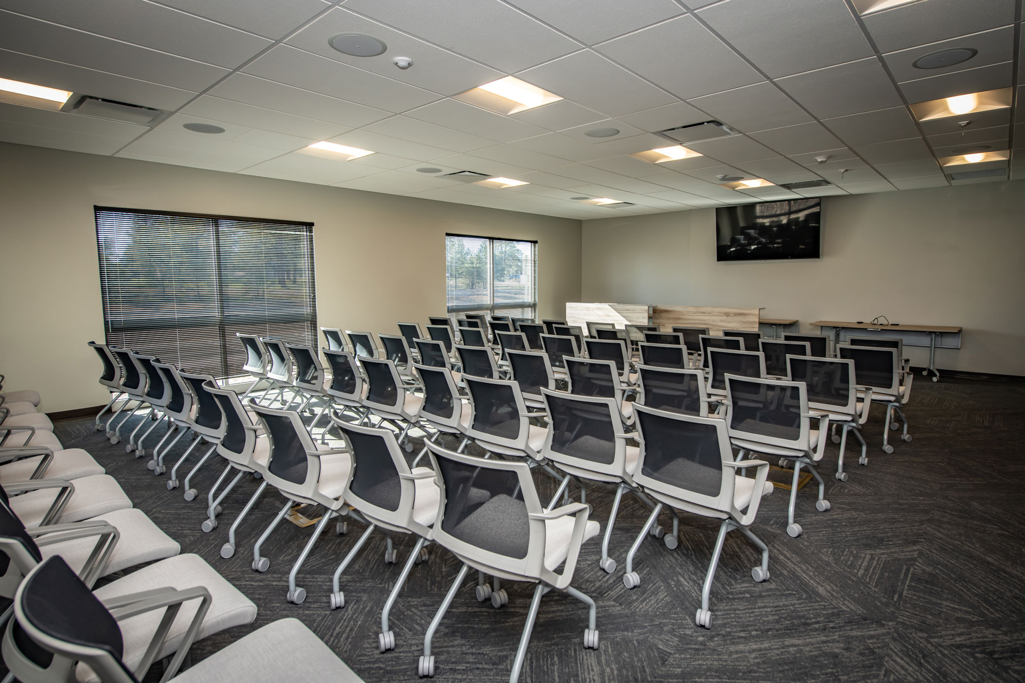 Conference room setup with rows of black and white chairs facing a large wall-mounted TV screen.