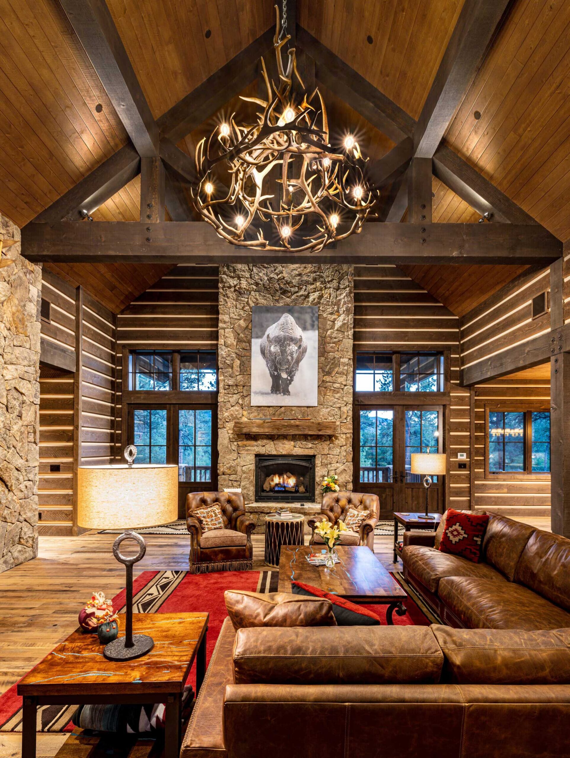 Cozy rustic living room featuring a stone fireplace, wooden beams, and a large antler chandelier. Co.